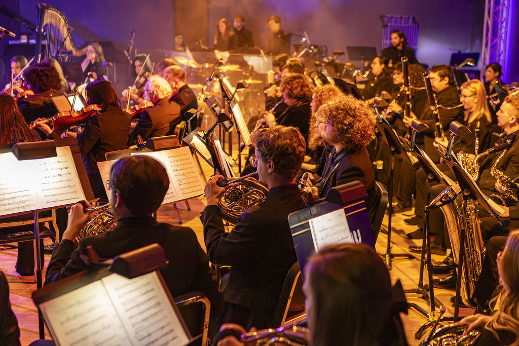 The WT symphony orchestra from stage left. The last row of string players, the brass, and some woodwinds are lit in a golden orange glow